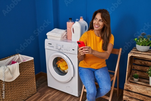 Young latin woman using smartphone waiting for washing machine at laundry room