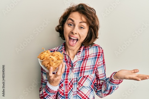 Young brunette woman holding bowl with italian pasta celebrating achievement with happy smile and winner expression with raised hand