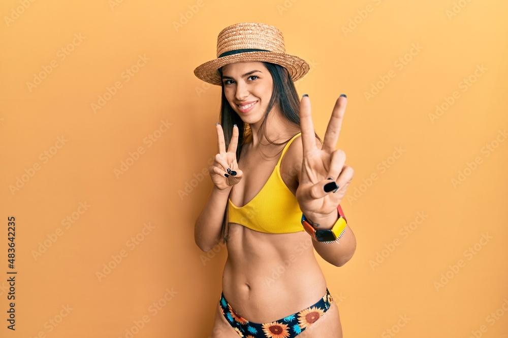 Young hispanic woman wearing bikini and summer hat smiling looking to the camera showing fingers doing victory sign. number two. Young hispanic woman wearing bikini and summer hat smiling looking to the camera showing fingers doing victory sign. number two.