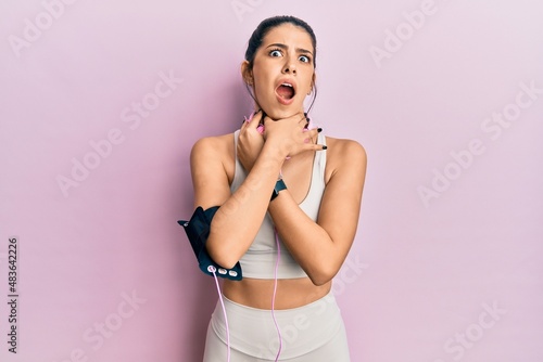 Young hispanic woman wearing gym clothes and using headphones shouting suffocate because painful strangle. health problem. asphyxiate and suicide concept.
