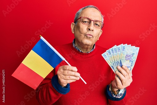 Handsome senior man with grey hair holding romania flag and leu banknotes looking at the camera blowing a kiss being lovely and sexy. love expression.