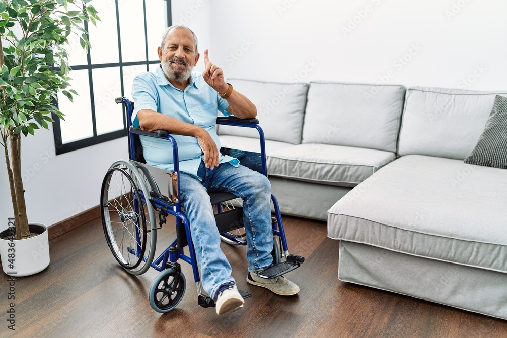 Handsome senior man sitting on wheelchair at the living room showing and pointing up with finger number one while smiling confident and happy. Handsome senior man sitting on wheelchair at the living room showing and pointing up with finger number one while smiling confident and happy.