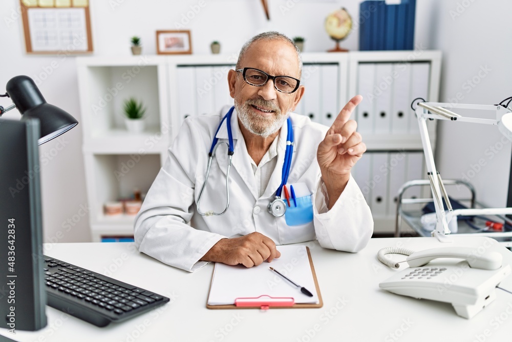 Mature doctor man at the clinic with a big smile on face, pointing with hand and finger to the side looking at the camera. Mature doctor man at the clinic with a big smile on face, pointing with hand and finger to the side looking at the camera.