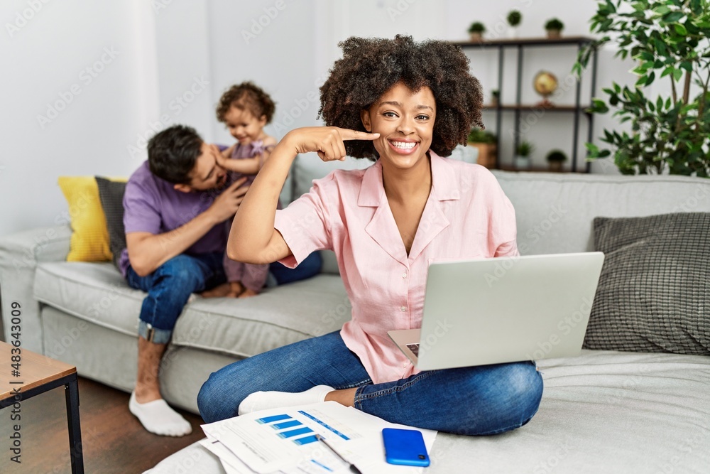 Mother of interracial family working using computer laptop at home smiling cheerful showing and pointing with fingers teeth and mouth. dental health concept.