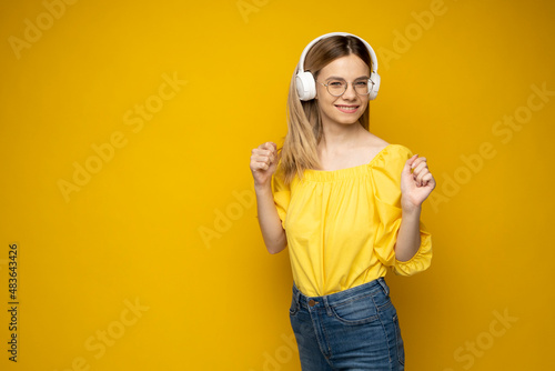 Beautiful attractive young blond woman wearing yellow t-shirt and glasses in white headphones listening music, dancing and laughing on yellow background in studio. Lifestyle.