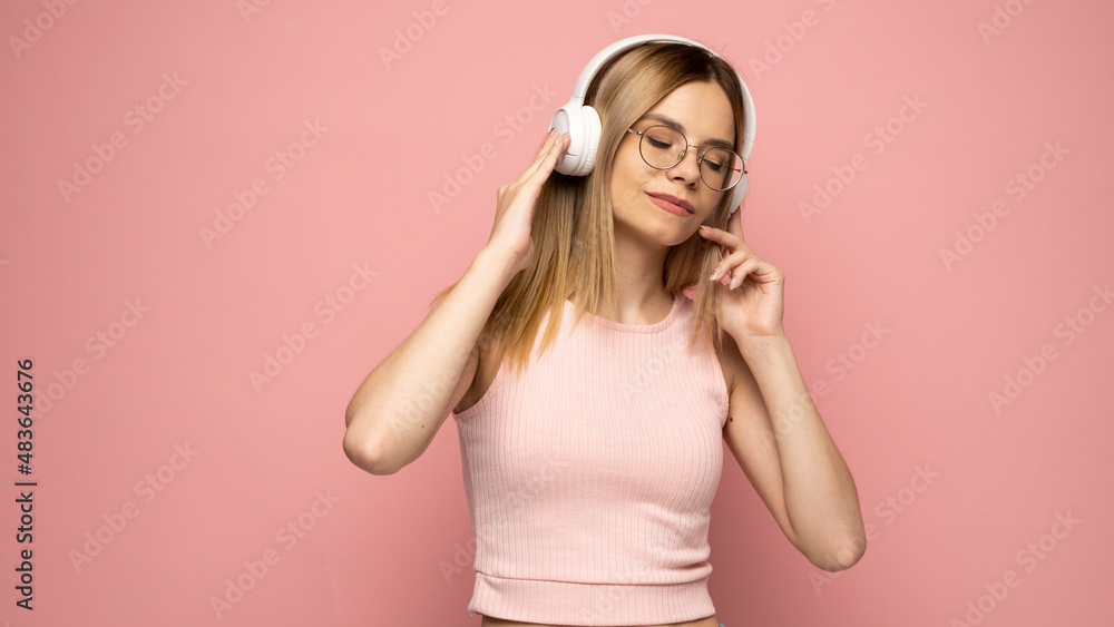 Beautiful attractive young blond woman wearing pink t-shirt and glasses in white headphones listening music on pink background in studio. Lifestyle. Beautiful attractive young blond woman wearing pink t-shirt and glasses in white headphones listening music on pink background in studio. Lifestyle.
