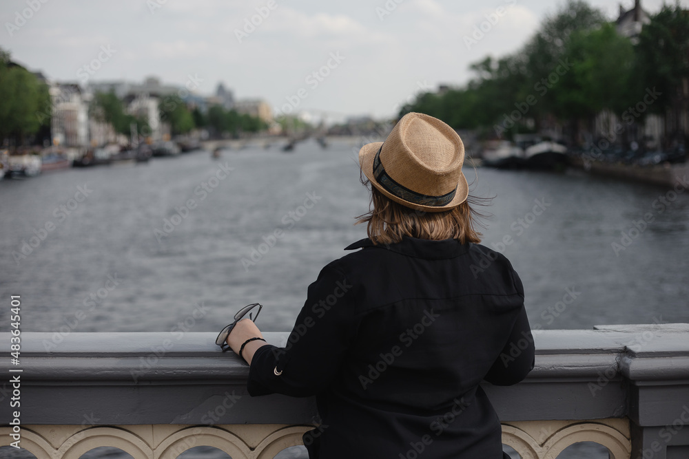 Obraz premium A girl leaning on railing looking at river in Amsterdam city on sunny day. Woman enjoying urban views with bridge on background. 