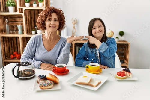 Family of mother and down syndrome daughter sitting at home eating breakfast amazed and smiling to the camera while presenting with hand and pointing with finger.