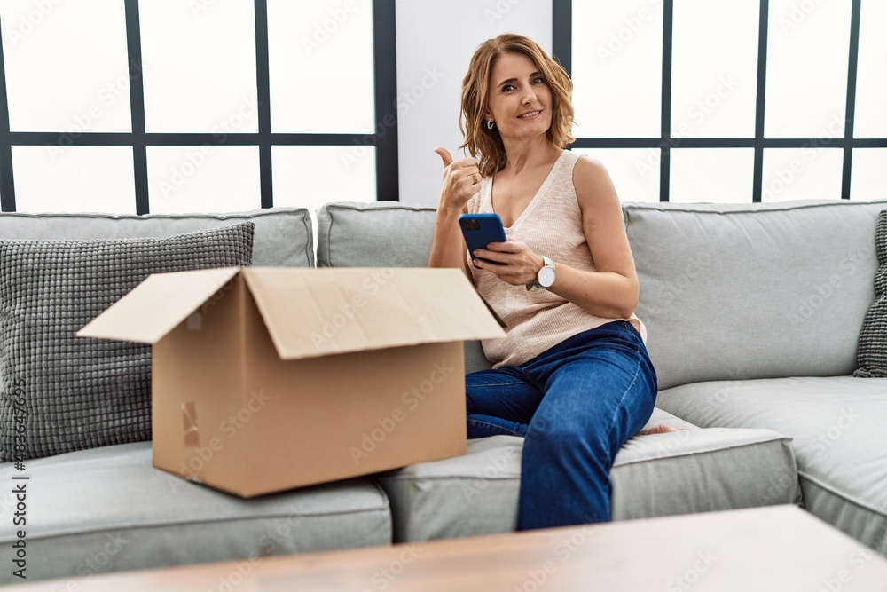 Middle age woman sitting on the sofa checking delivery package on the phone smiling happy and positive, thumb up doing excellent and approval sign Middle age woman sitting on the sofa checking delivery package on the phone smiling happy and positive, thumb up doing excellent and approval sign