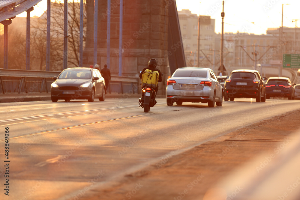 Kurier na skuterze jedzie mostem Grunwaldzkim we Wrocławiu, pod zachodzące słońce. - obrazy, fototapety, plakaty 
