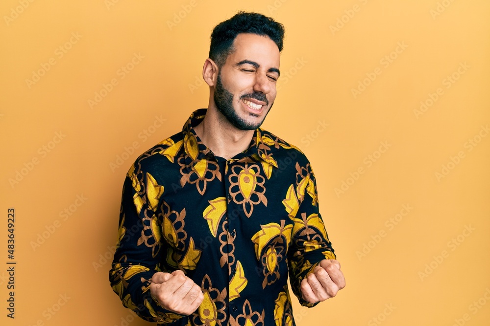 Young hispanic man wearing casual shirt very happy and excited doing winner gesture with arms raised, smiling and screaming for success. celebration concept. Young hispanic man wearing casual shirt very happy and excited doing winner gesture with arms raised, smiling and screaming for success. celebration concept.