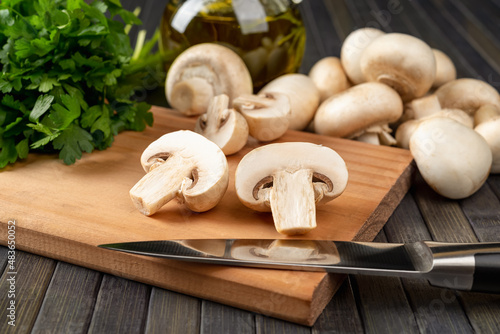 Raw champignon cut in half and and kitchen knife on a cutting board, whole button mushrooms and bunch of parsley over dark background. Vegetarian food ingredient, vegetable protein.