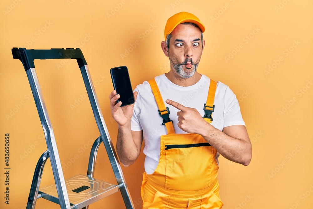 Handsome middle age man with grey hair standing by ladder showing smartphone making fish face with mouth and squinting eyes, crazy and comical. Handsome middle age man with grey hair standing by ladder showing smartphone making fish face with mouth and squinting eyes, crazy and comical.