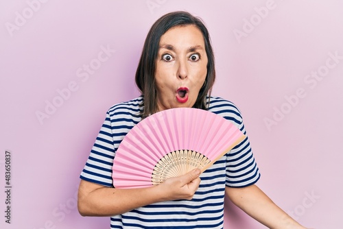 Middle age hispanic woman waving hand fan cooling air in summer scared and amazed with open mouth for surprise, disbelief face © Krakenimages.com