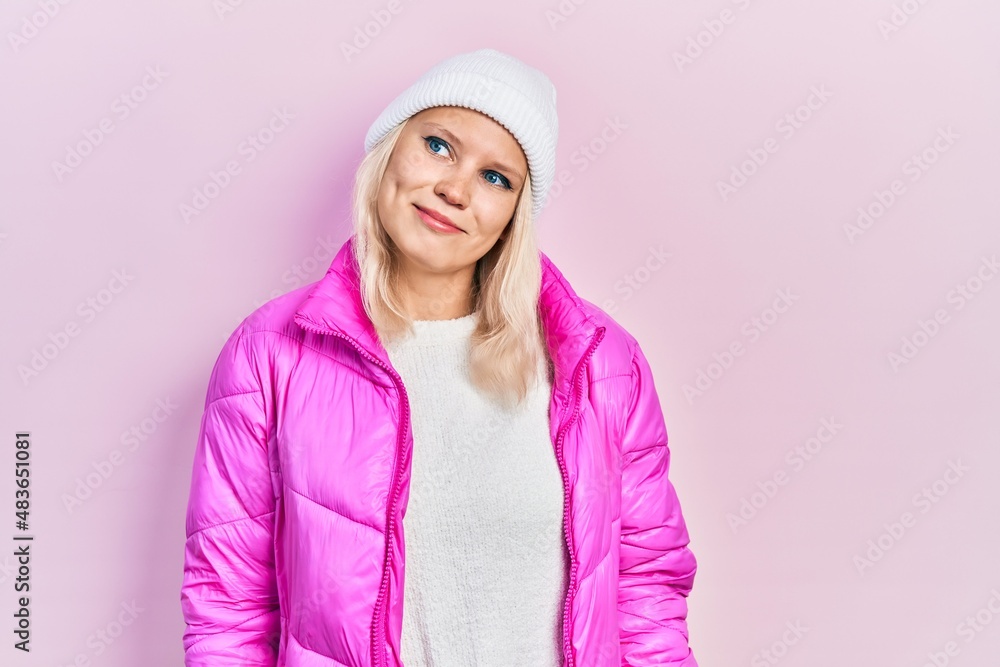Beautiful caucasian blonde woman wearing wool hat and winter coat looking away to side with smile on face, natural expression. laughing confident. Beautiful caucasian blonde woman wearing wool hat and winter coat looking away to side with smile on face, natural expression. laughing confident.