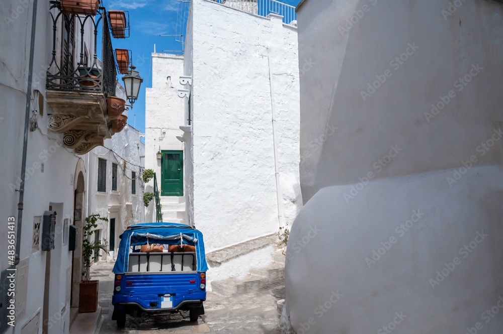 Ostuni, Puglia, Italy. August 2021. Amazing view on one of the white alleys of the old town. A blue-colored tourist truck is in the middle of the alley.