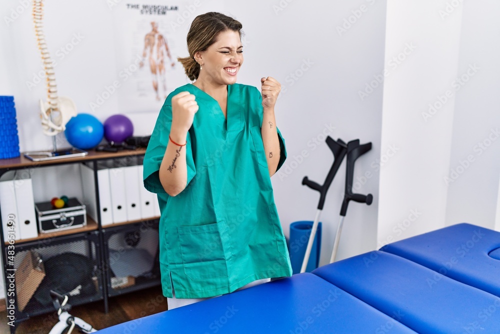 Young hispanic woman wearing physiotherapist uniform standing at clinic very happy and excited doing winner gesture with arms raised, smiling and screaming for success. celebration concept. Young hispanic woman wearing physiotherapist uniform standing at clinic very happy and excited doing winner gesture with arms raised, smiling and screaming for success. celebration concept.