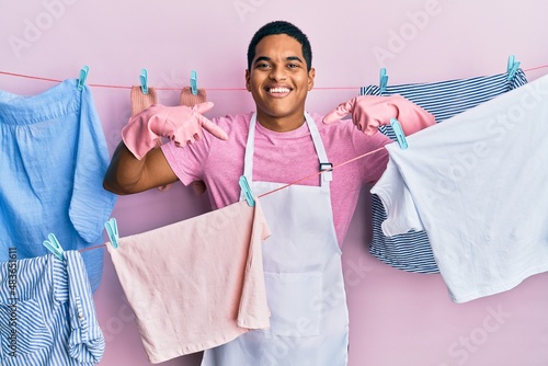 Young handsome hispanic man wearing cleaner apron holding clothes on clothesline looking confident with smile on face, pointing oneself with fingers proud and happy.