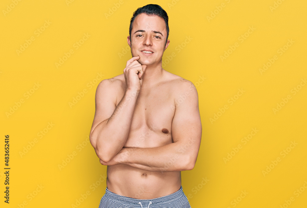Fototapeta premium Handsome young man wearing swimwear shirtless looking confident at the camera smiling with crossed arms and hand raised on chin. thinking positive.