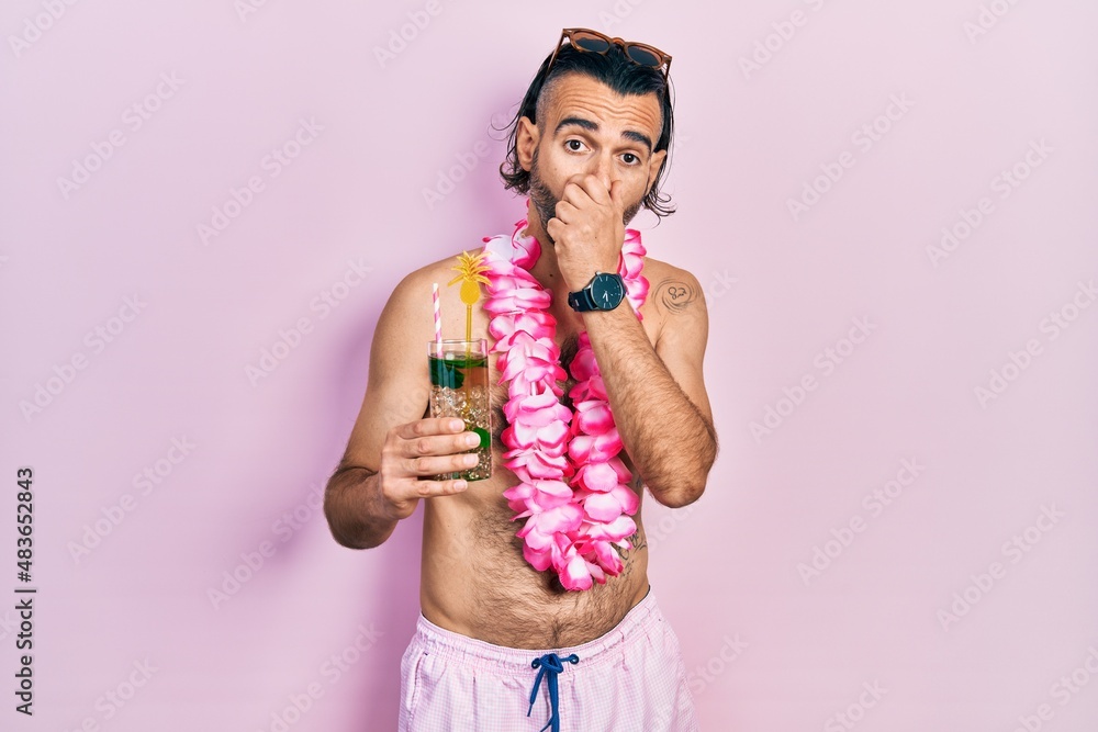 Young hispanic man wearing swimsuit and hawaiian lei drinking tropical cocktail shocked covering mouth with hands for mistake. secret concept. Young hispanic man wearing swimsuit and hawaiian lei drinking tropical cocktail shocked covering mouth with hands for mistake. secret concept.