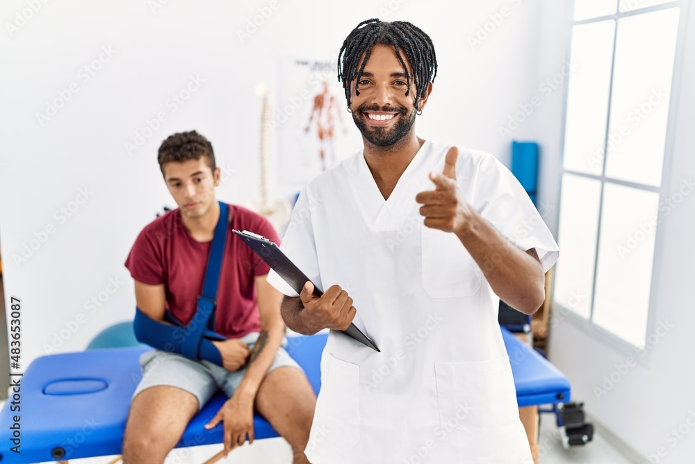 Obraz premium Young hispanic man working at pain recovery clinic with a man with broken arm pointing fingers to camera with happy and funny face. good energy and vibes.