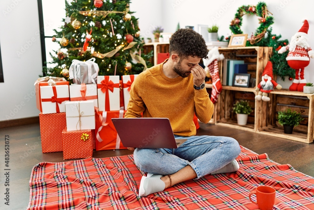 Arab young man using laptop sitting by christmas tree tired rubbing nose and eyes feeling fatigue and headache. stress and frustration concept. Arab young man using laptop sitting by christmas tree tired rubbing nose and eyes feeling fatigue and headache. stress and frustration concept.