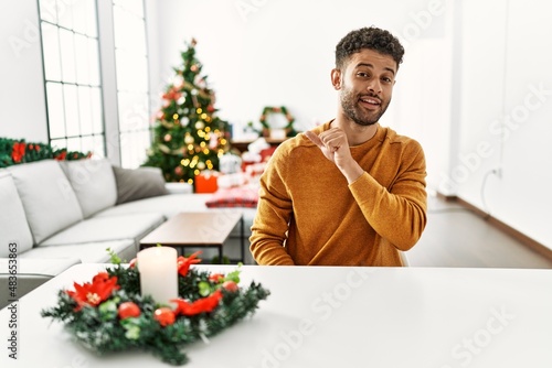 Arab young man sitting on the table by christmas tree smiling with happy face looking and pointing to the side with thumb up.