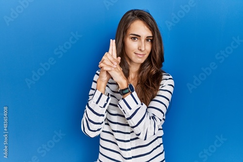 Young hispanic woman standing over blue isolated background holding symbolic gun with hand gesture, playing killing shooting weapons, angry face