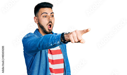 Young hispanic man with beard wearing casual denim jacket pointing with finger surprised ahead, open mouth amazed expression, something on the front