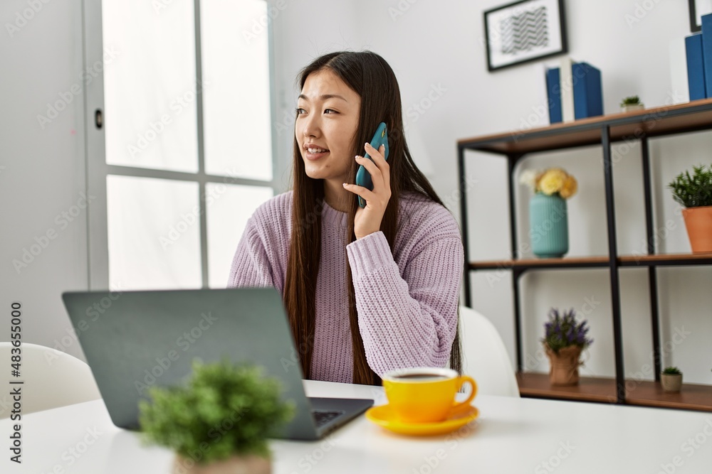 Young chinese girl using laptop and talking on the smartphone sitting on the table at home. Young chinese girl using laptop and talking on the smartphone sitting on the table at home.