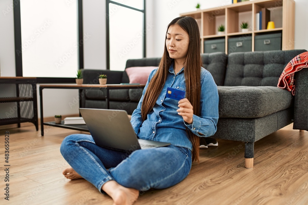 Young chinese girl using laptop and credit card sitting on the floor at home.