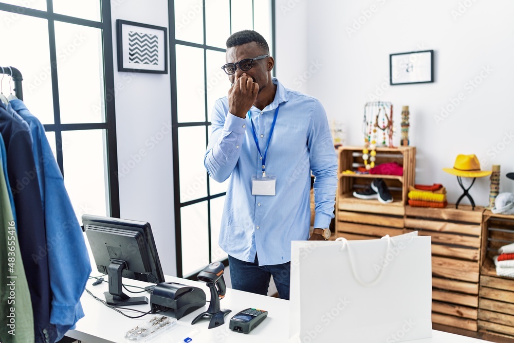 Young african man working as manager at retail boutique smelling something stinky and disgusting, intolerable smell, holding breath with fingers on nose. bad smell - obrazy, fototapety, plakaty 