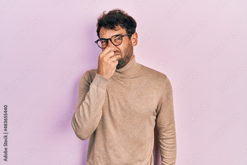 Handsome man with beard wearing turtleneck sweater and glasses smelling something stinky and disgusting, intolerable smell, holding breath with fingers on nose. bad smell Handsome man with beard wearing turtleneck sweater and glasses smelling something stinky and disgusting, intolerable smell, holding breath with fingers on nose. bad smell