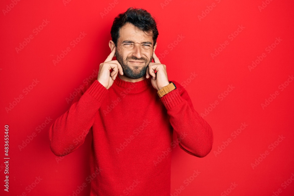 Handsome man with beard wearing casual red sweater covering ears with fingers with annoyed expression for the noise of loud music. deaf concept. Handsome man with beard wearing casual red sweater covering ears with fingers with annoyed expression for the noise of loud music. deaf concept.