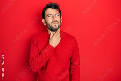 Handsome man with beard wearing casual red sweater touching painful neck, sore throat for flu, clod and infection