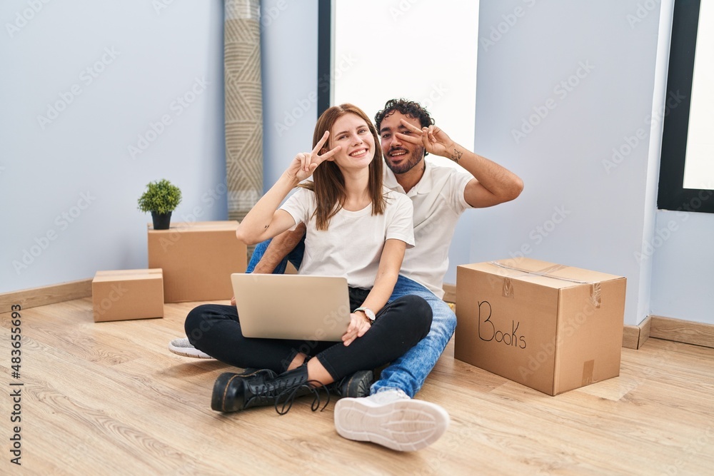 Young couple using laptop at new home doing peace symbol with fingers over face, smiling cheerful showing victory Young couple using laptop at new home doing peace symbol with fingers over face, smiling cheerful showing victory