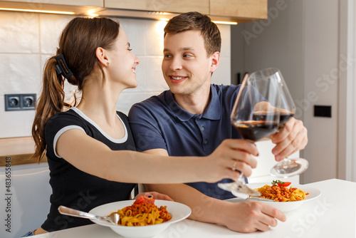 Valentine day romantic dinner at home. Happy couple smiling, clinking glasses and eating pasta with wine at kitchen. Concept of domestic lifestyle, love, happy marriage and togetherness.