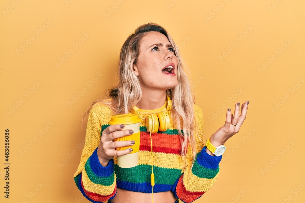 Beautiful young blonde woman drinking cup of coffee wearing headphones crazy and mad shouting and yelling with aggressive expression and arms raised. frustration concept. Beautiful young blonde woman drinking cup of coffee wearing headphones crazy and mad shouting and yelling with aggressive expression and arms raised. frustration concept.