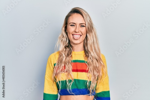 Beautiful young blonde woman wearing colored sweater sticking tongue out happy with funny expression. emotion concept.