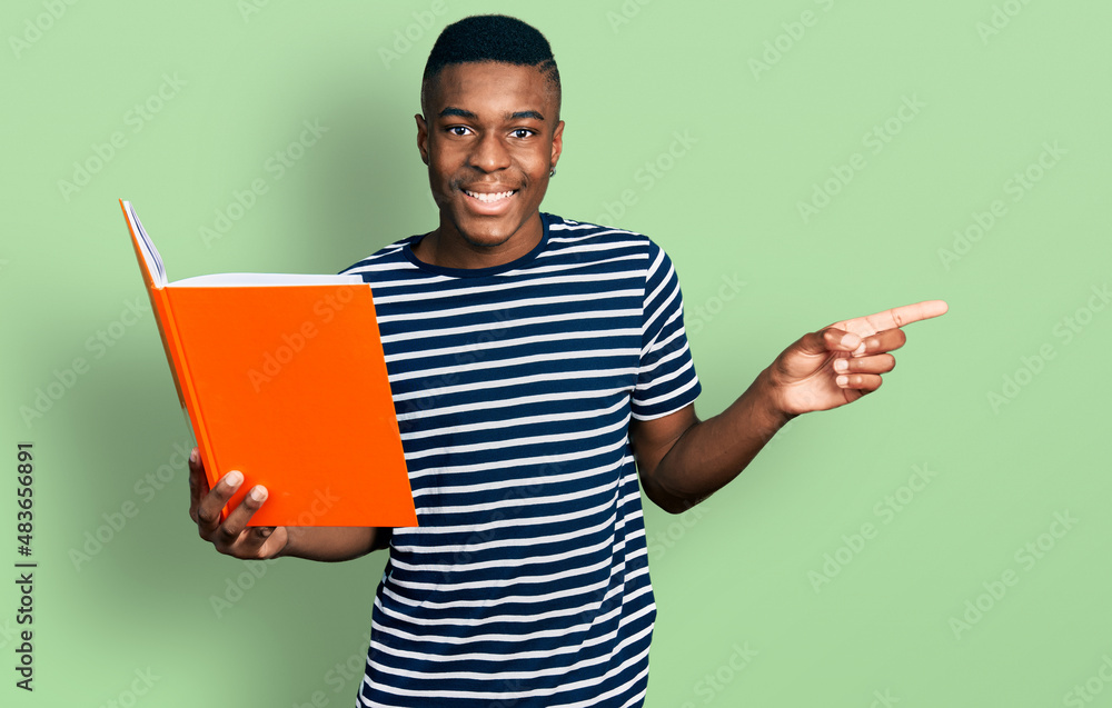 Young african american man holding book smiling happy pointing with hand and finger to the side Young african american man holding book smiling happy pointing with hand and finger to the side