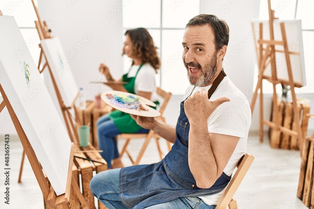 Hispanic middle age man and mature woman at art studio pointing thumb up to the side smiling happy with open mouth Hispanic middle age man and mature woman at art studio pointing thumb up to the side smiling happy with open mouth
