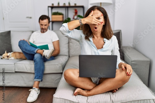 Hispanic middle age couple at home, woman using laptop peeking in shock covering face and eyes with hand, looking through fingers with embarrassed expression.