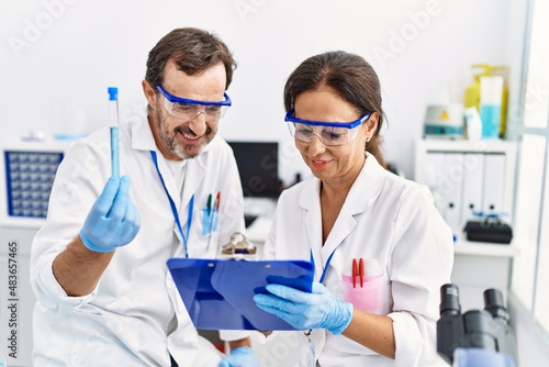 Middle age man and woman partners wearing scientist uniform writing on clipboard looking test tube at laboratory