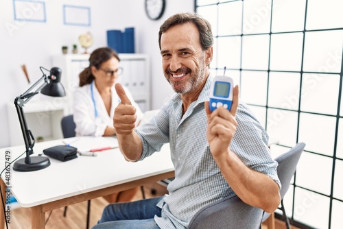 Middle age man at doctor clinic holding glucose meter device smiling happy and positive, thumb up doing excellent and approval sign