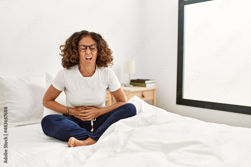 Middle age hispanic woman sitting on the bed at home with hand on stomach because indigestion, painful illness feeling unwell. ache concept. Middle age hispanic woman sitting on the bed at home with hand on stomach because indigestion, painful illness feeling unwell. ache concept.