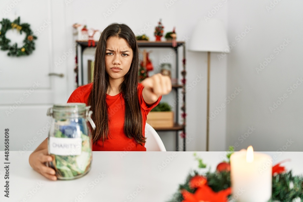Young brunette woman on christmas holding charity jar with money at home pointing with finger to the camera and to you, confident gesture looking serious