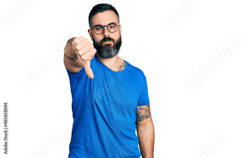 Hispanic man with beard wearing casual t shirt and glasses looking unhappy and angry showing rejection and negative with thumbs down gesture. bad expression.