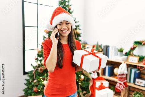 Young latin woman talking on the smartphone standing by christmas tree at home