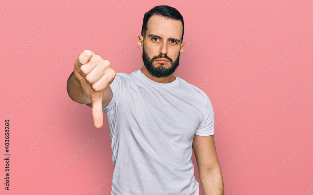Young man with beard wearing casual white t shirt looking unhappy and angry showing rejection and negative with thumbs down gesture. bad expression. Young man with beard wearing casual white t shirt looking unhappy and angry showing rejection and negative with thumbs down gesture. bad expression.