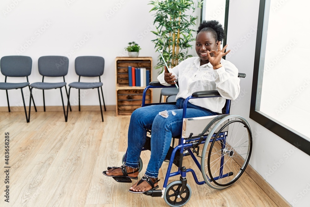 Young black woman sitting on wheelchair at waiting room smiling positive doing ok sign with hand and fingers. successful expression. Young black woman sitting on wheelchair at waiting room smiling positive doing ok sign with hand and fingers. successful expression.